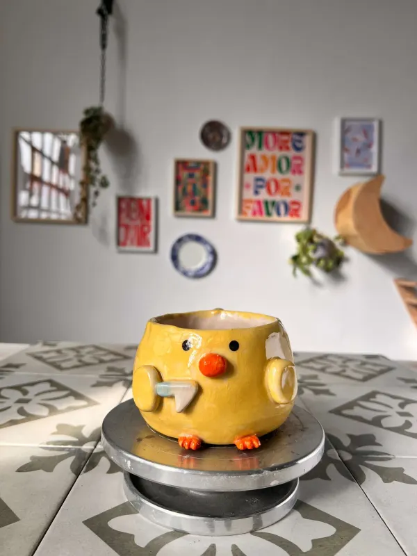 Macetero de cerámica con forma de pollito, esmaltado en amarillo con el pico y las patas naranjas, sobre una torneta